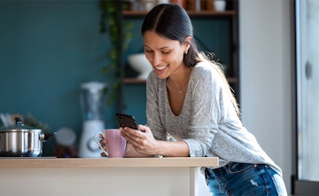 Eine Frau schaut am Küchentisch auf ihr Handy und lächelt.